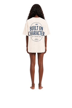 A woman shown from the back wearing an oversized cream-colored t-shirt with a navy blue graphic print that reads 'BUILT ON CHARACTER' inside a circle, on a solid white background.