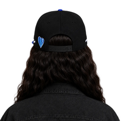 Back view of a person with long wavy dark hair wearing a black baseball cap with blue accents and a blue heart logo on the side, paired with a black denim jacket against a light gray background.