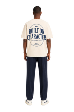 A man viewed from the back wearing a cream-colored t-shirt with a large navy blue graphic on the back that reads 'BUILT ON CHARACTER', paired with navy blue pants and white sneakers, against a plain white background.